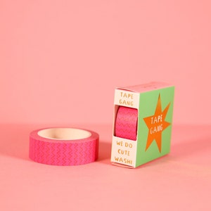 Puede incluir: Un rollo de cinta washi con estampado rosa y blanco con una caja verde y dorada que dice "Tape Gang" y "We do cute washi".