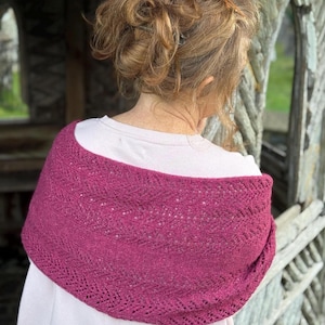 May include: A knitted shawl in a deep magenta colour, draped over a person's shoulders. The shawl features a delicate openwork pattern and is made of soft yarn. The person is wearing a white shirt. The background is blurred.