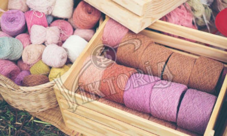 May include: A collection of colorful yarn balls in a woven basket and wooden crate. The yarn colors include pink, purple, brown, and white. The yarn is likely used for knitting or crocheting projects.