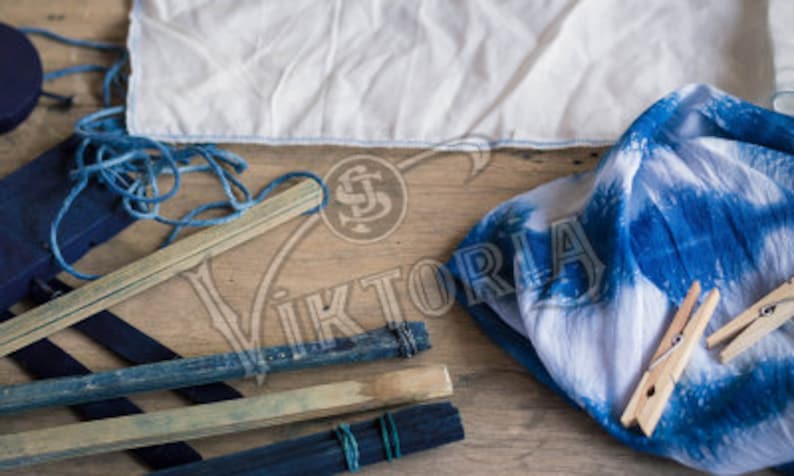 May include: An overhead shot of various indigo dyeing supplies. Includes a white cloth, blue-dyed fabric, wooden sticks, string, and clothespins. The items are arranged on a wooden surface, showcasing the process of creating tie-dye textiles.
