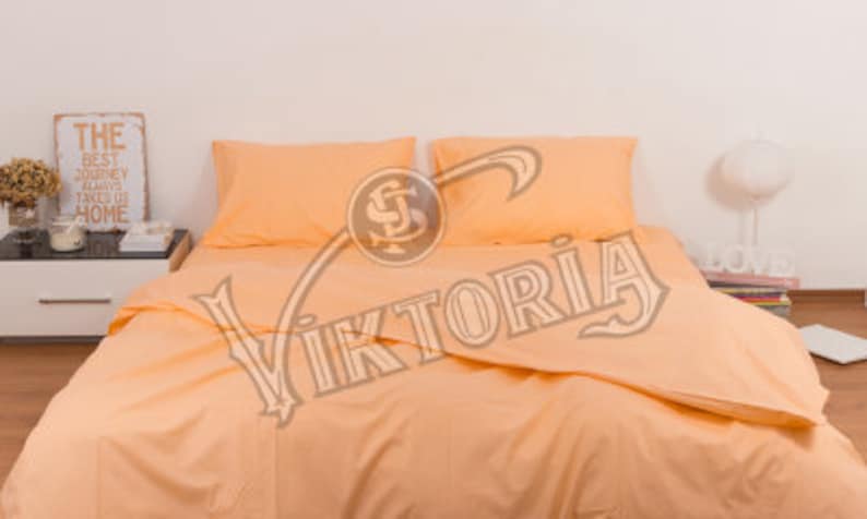May include: Peach-colored bed linens set on a bed. The set includes a duvet cover and two pillowcases. A nightstand with a decorative sign that reads "THE BEST JOURNEY ALWAYS TAKES US HOME" is visible in the background.