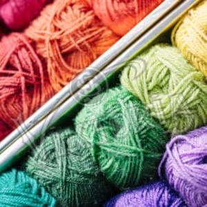 May include: A close-up of colorful yarn balls in shades of red, orange, green, yellow, and purple. Two silver knitting needles are placed diagonally across the yarn balls, suggesting a crafting theme. The yarn is tightly wound, and the image is well-lit.