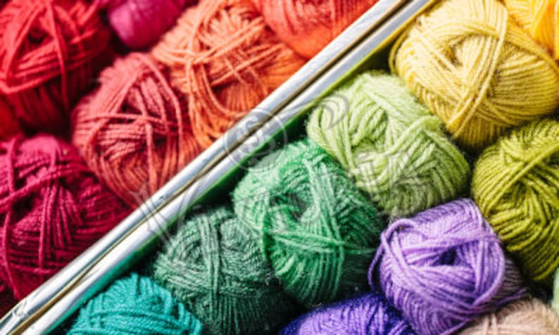 May include: A close-up of colorful yarn balls in shades of red, orange, green, yellow, and purple. Two silver knitting needles are placed diagonally across the yarn balls, suggesting a crafting theme. The yarn is tightly wound, and the image is well-lit.