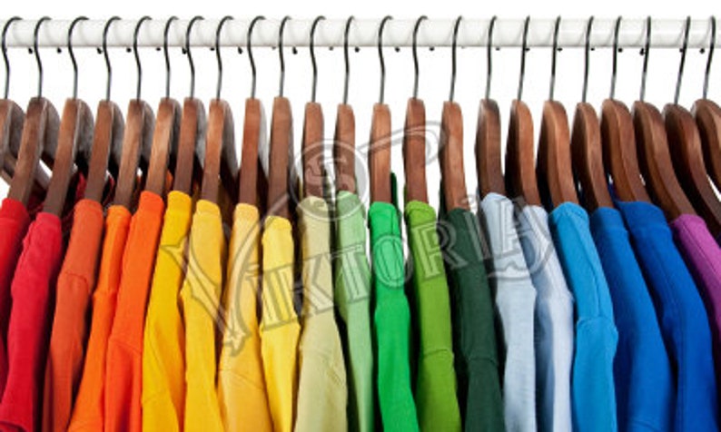 May include: A row of colorful t-shirts hanging on wooden hangers. The shirts are arranged in a rainbow spectrum, from red to purple. The hangers are suspended from a white rod with black hooks, creating a visually appealing display.