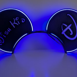 Puede incluir: Orejas de ratón negras con un contorno LED azul y un brillo azul a juego. La oreja izquierda tiene el texto "Wish Kid" en escritura blanca, y la oreja derecha presenta un logotipo blanco.