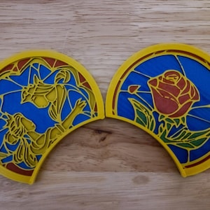 May include: Two yellow, semi-circular decorations with stained-glass designs. One depicts a woman and a beast, the other a red rose. The designs are set against a blue background with yellow and red accents.