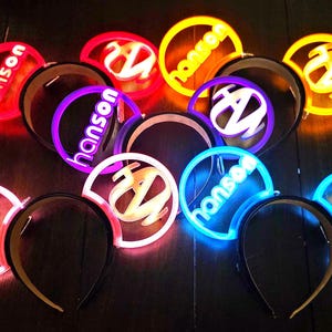 Op de afbeelding: Een verzameling verlichte haarbanden in verschillende kleuren, waaronder rood, paars, geel en blauw. Elke haarband heeft het woord "hanson" en een gestileerd "Z"-logo, allemaal verlicht met neon-achtige effecten. De haarbanden staan tegen een donkere achtergrond.