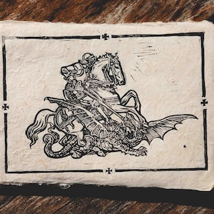 May include: A monochrome linocut print showing a knight on horseback battling a dragon. The artwork is on textured paper with a black border and cross designs at the corners. The print is set against a wooden background.