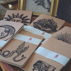 Peut inclure: Plusieurs carnets en papier kraft avec des illustrations à l'encre noire. Les motifs incluent un soleil, une lune, un œil, un dragon et un taureau. Les carnets sont liés par des bandes de papier blanc. Un dessin encadré et une figurine de serpent en laiton sont également visibles.