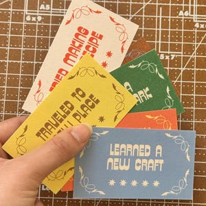 May include: A hand holding a selection of colourful rectangular cards. Each card displays text in a retro font, including "Learned a New Craft" and "Traveled to a New Place." The cards have decorative borders and star accents.