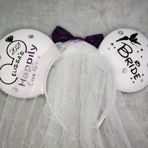 May include: White Minnie Mouse ears with a white tulle veil. One ear has the text "Happily Ever After" and the date "2021" with the name "Elissa's". The other ear has the text "To Be Bride" with a silhouette of a fairy.