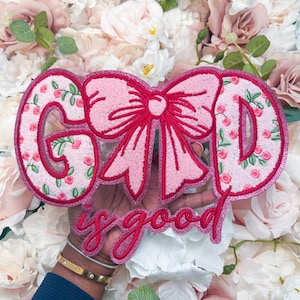 May include: A decorative patch featuring the text "GOD is good" with a large pink bow replacing the letter O. The letters G, D, and the bow are filled with a light pink fabric patterned with small red roses and green leaves, all outlined in a thick, sparkling dark pink border.