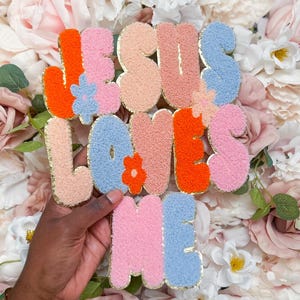 May include: A tufted wall hanging featuring the text JESUS LOVES ME in a multi-colored palette of orange, light pink, peach, and light blue. The letters are accented with small flower shapes and have a gold-toned metallic border.