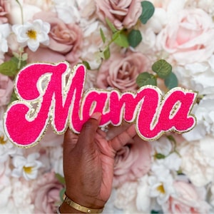 Puede incluir: Un parche de color rosa intenso con la palabra Mama en letra cursiva. Las letras tienen una superficie texturizada y difusa, delineadas con un borde de purpurina dorada brillante, creando un accesorio decorativo llamativo.