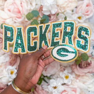 May include: A decorative patch featuring the word PACKERS in green sequins with a gold border, accompanied by a green and white sequined G logo. The letters and logo are outlined in gold, creating a bold, textured appearance for apparel.