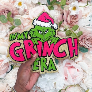 Op de afbeelding: Een chenille patch met een groene Grinch-figuur die een roze kerstmuts met witte rand draagt. De patch bevat de tekst "IN MY GRINCH ERA" in felroze en groene letters met een gouden glitterrand en zwarte omlijningen.