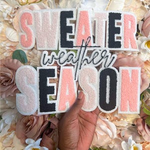 May include: A decorative sign featuring the text SWEATER weather SEASON in a mix of pink, black, and white colors. The letters are crafted with a textured, fuzzy material and outlined with white borders, creating a layered, dimensional appearance.