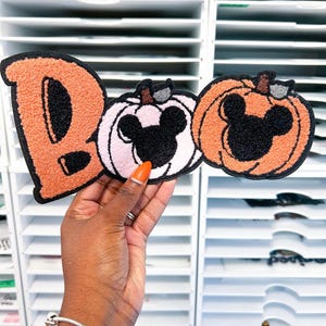 May include: A chenille patch featuring the word BOO, where the letter B is orange, the letter O is a light pink pumpkin, and the second O is an orange pumpkin. Both pumpkins feature a black mouse-ear silhouette in the center.