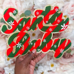 Puede incluir: Un letrero decorativo con el texto Santa Baby en letra cursiva. Las letras tienen un patrón de rayas diagonales en rojo y verde con pequeños copos de nieve blancos. Toda la pieza está delineada con un borde de purpurina dorada.