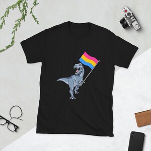Pode incluir: Camiseta preta com um dinossauro cinza segurando uma bandeira do orgulho pansexual. A bandeira é amarela, azul e rosa.