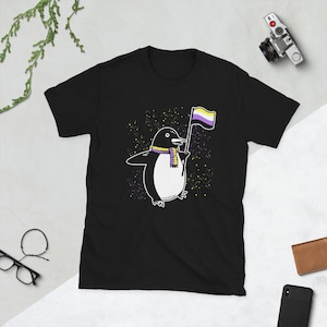 May include: Black t-shirt featuring a cartoon penguin wearing a yellow and purple scarf and holding a yellow, purple and white flag. The background is a black and white starry pattern.