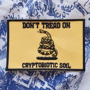 May include: A yellow patch with a black border and black text that reads "DON'T TREAD ON CRYPTOBIOTIC SOIL". A black and yellow coiled snake is in the center of the patch.