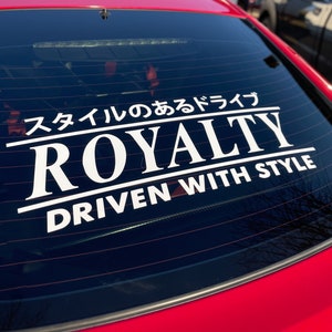 May include: A white vinyl sticker on the back window of a red car. The sticker says "ROYALTY" in large white letters with a smaller line of text that says "DRIVEN WITH STYLE" below it. Above the word "ROYALTY" is a line of Japanese text that says "スタイルのあるドライブ".
