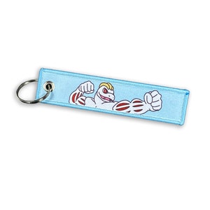 May include: A light blue key chain featuring a cartoon image of a white, muscular creature with red veins and a yellow mohawk. The creature is flexing its biceps.