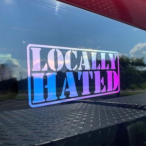 Op de afbeelding: Een sticker op een autoruit met de tekst "Lokaal gehaat" in wit met een blauwe en paarse iriserende achtergrond.