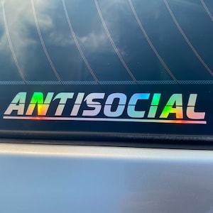 Puede incluir: Una pegatina para la ventana trasera de un coche con la palabra "ANTISOCIAL" en blanco, en negrita, con un efecto arcoíris iridiscente.