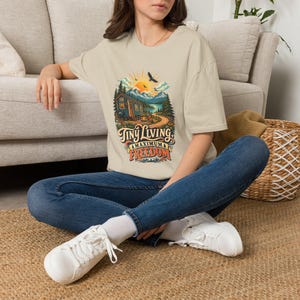 May include: A beige short-sleeve t-shirt featuring a colorful graphic of a small cabin in a mountain landscape with a sun, trees, and the text Tiny Living, Maximum Freedom. The shirt is paired with blue denim jeans and white sneakers.