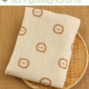 May include: Cream-colored baby blanket with a repeating pattern of brown lion faces. The blanket is made of a soft, textured fabric and is folded on a woven tray. The text "SpringdesignCrafts" is visible in the background.