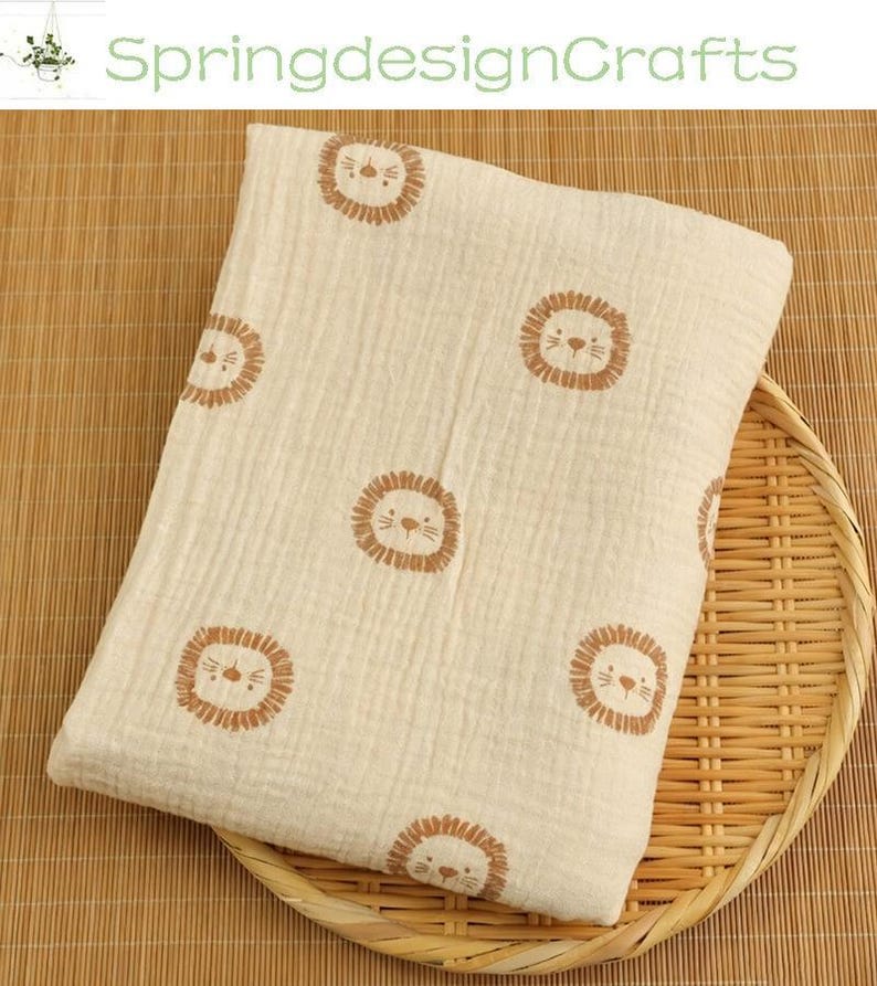 May include: Cream-colored baby blanket with a repeating pattern of brown lion faces. The blanket is made of a soft, textured fabric and is folded on a woven tray. The text "SpringdesignCrafts" is visible in the background.