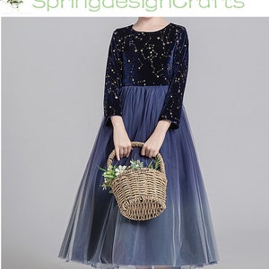May include: A navy blue dress with a celestial pattern on the long sleeves and bodice. The dress features a tulle skirt that fades from navy to a lighter blue. A woven basket with flowers is held in front of the dress.