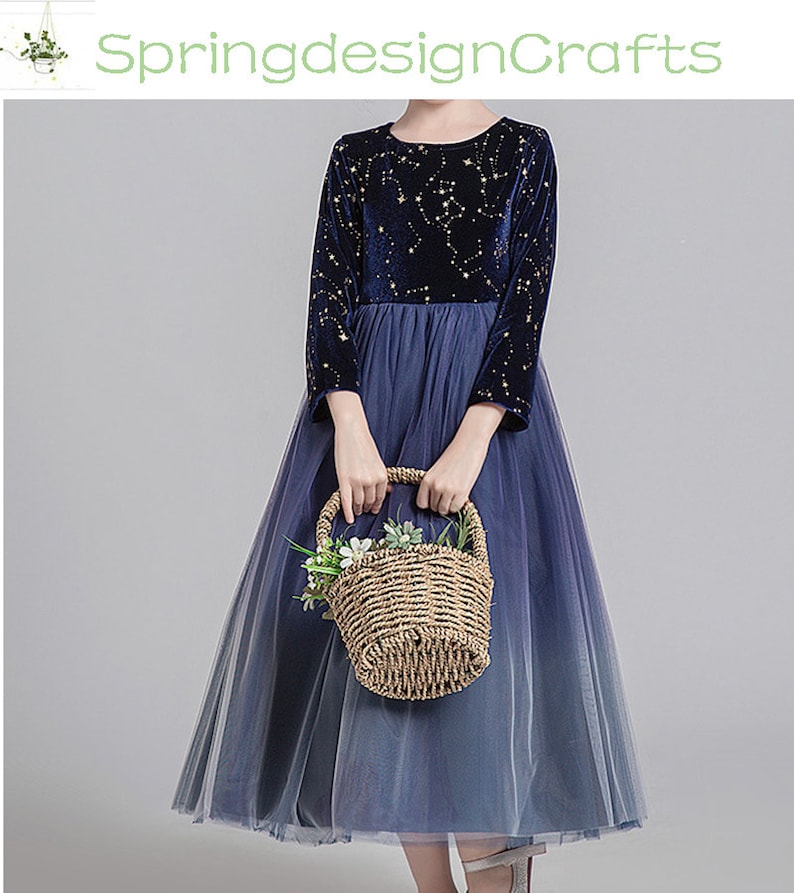 May include: A navy blue dress with a celestial pattern on the long sleeves and bodice. The dress features a tulle skirt that fades from navy to a lighter blue. A woven basket with flowers is held in front of the dress.