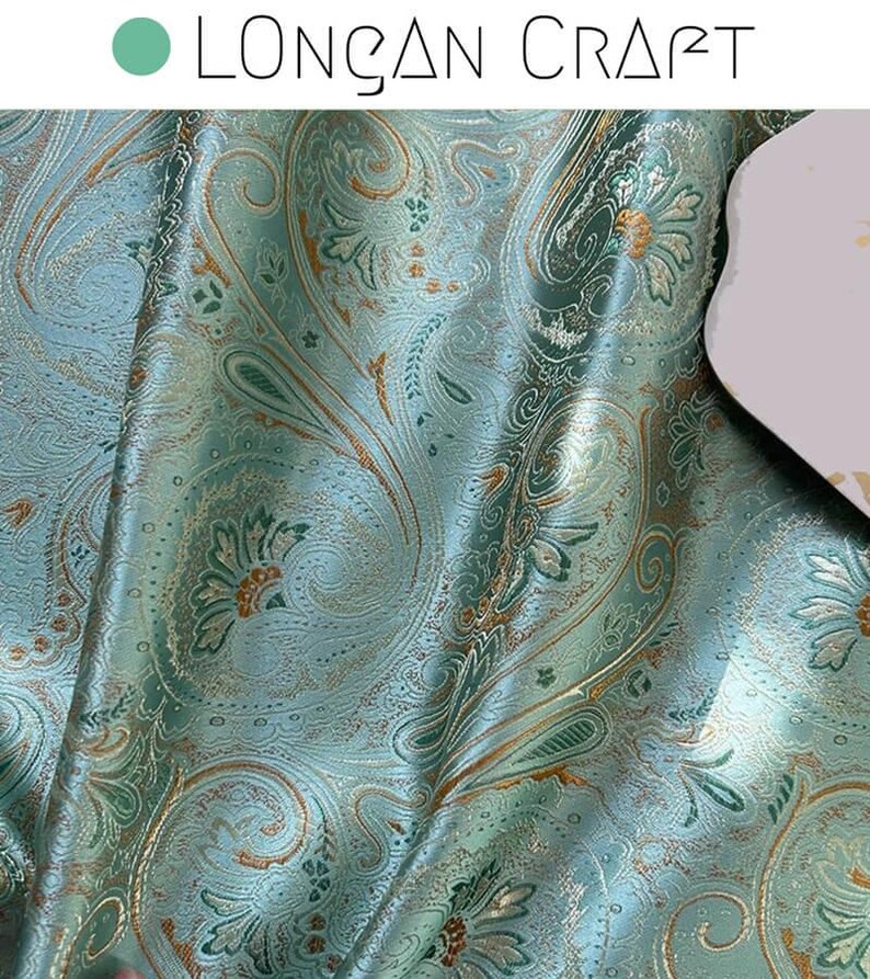 May include: Teal fabric with a paisley pattern in gold and green. The fabric has a subtle sheen and a textured surface.