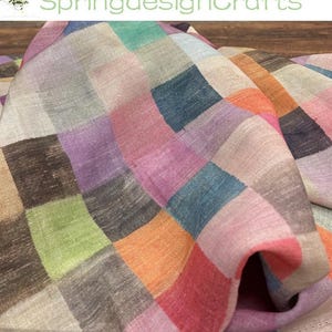 May include: A woven fabric featuring a multicolored checkered pattern with squares in shades of orange, pink, purple, blue, green, and beige. The textile has a visible textured weave and is draped to show folds and depth across the surface.