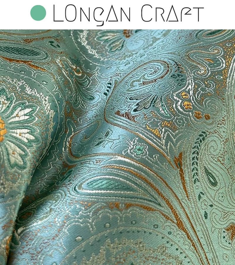 May include: Close-up of a teal fabric with a paisley pattern in gold and green. The fabric has a subtle sheen and a textured surface.
