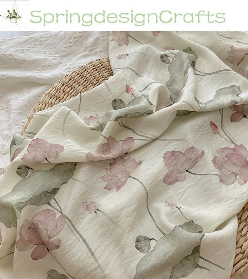 Peut inclure: Tissu blanc cass&eacute; avec un motif floral d&eacute;licat de fleurs de lotus roses et de feuilles vertes. Le tissu est drap&eacute; sur un panier en osier, avec le texte "SpringdesignCrafts" en vert.