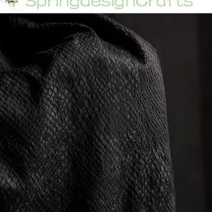 Op de afbeelding: Een stuk zwart materiaal met een getextureerd, herhalend schubachtig patroon over het oppervlak. De stof heeft een donkere, matte afwerking met subtiele variaties in tint, wat zorgt voor een uitgesproken, tactiele uitstraling over het materiaal.