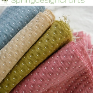 Op de afbeelding: Close-up van verschillende rollen stof in verschillende kleuren: lichtblauw, beige, olijfgroen en roze. Elke stof heeft een getextureerd oppervlak met kleine witte stippen. De tekst "SpringdesignCrafts" is rechtsboven zichtbaar.