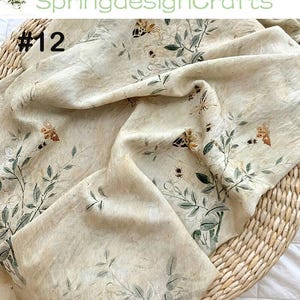 Peut inclure: Tissu cr&egrave;me avec un imprim&eacute; botanique repr&eacute;sentant des papillons, des feuilles et de petits insectes. Le tissu est drap&eacute; sur un panier en osier. Les textes "SpringdesignCrafts" et "#12" sont visibles.
