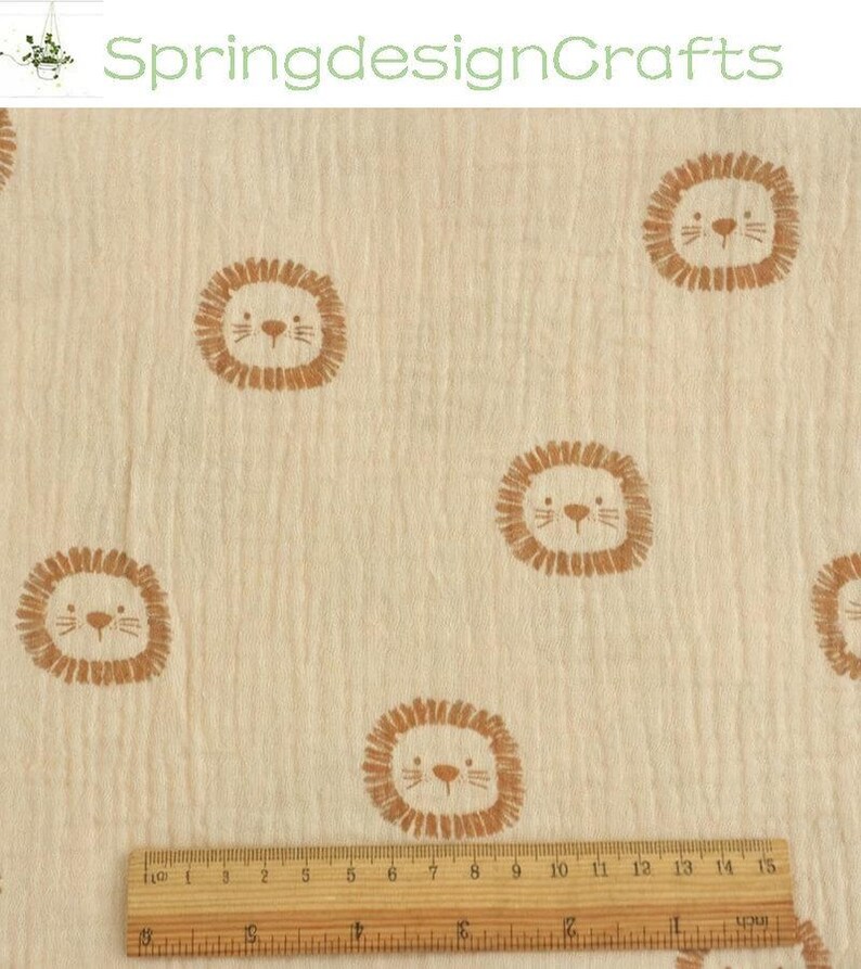 May include: Cream-colored fabric with a repeating pattern of cartoon lion faces in brown. A wooden ruler with inch markings is at the bottom. The text "SpringdesignCrafts" is at the top. The fabric appears to be muslin.