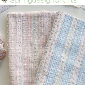 May include: Two fabric rolls, one pink and one blue, with a vertical striped pattern. The pink fabric has a gingham-like texture, while the blue fabric has a similar texture. Both fabrics feature floral and dotted accents.