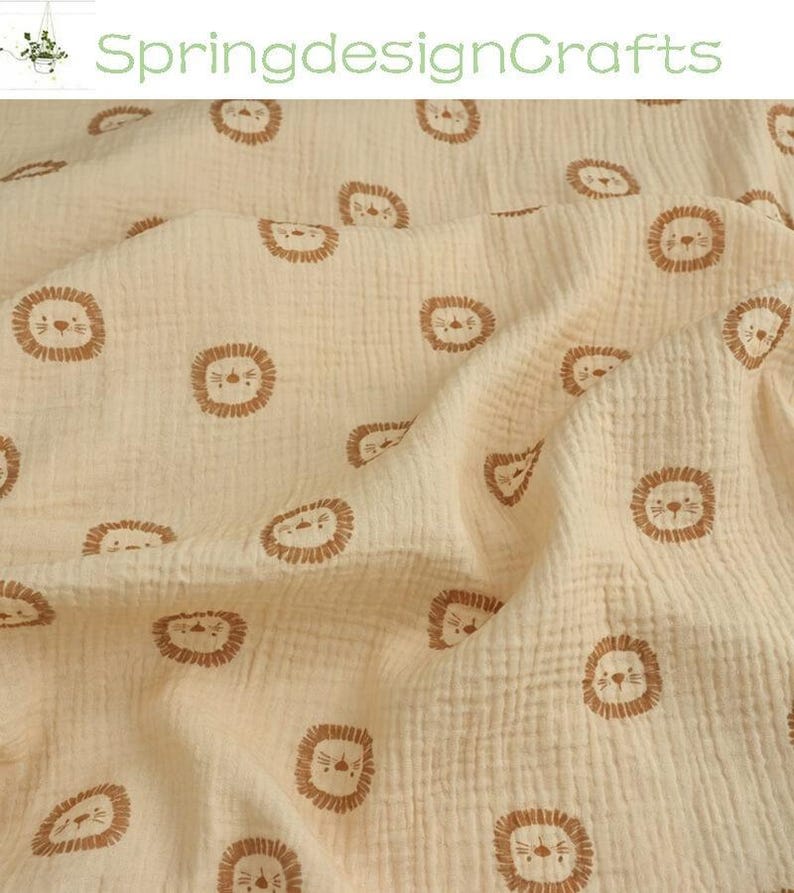 May include: Cream-colored fabric with a repeating pattern of brown lion faces. The fabric has a textured, crinkled appearance. The text "SpringdesignCrafts" is visible in the upper left corner. This fabric is suitable for baby blankets, swaddles, or other nursery items.