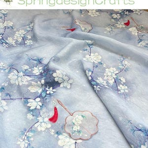 Pode incluir: Tecido azul claro com um padrão floral de flores brancas e galhos azuis. Um leque decorativo com uma borla vermelha é visível. O texto "SpringdesignCrafts" é visível.