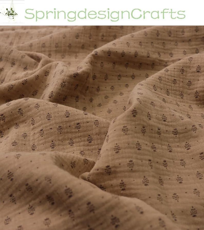 May include: Close-up of tan fabric with a repeating pattern of small, dark floral shapes. The fabric is soft and textured, with visible folds and shadows. The text "SpringdesignCrafts" is in green at the top.