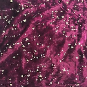 May include: Close-up of a deep burgundy velvet fabric with a constellation pattern. The design features small, gold-colored stars and connecting lines, creating a celestial theme. The fabric has a soft, textured appearance.