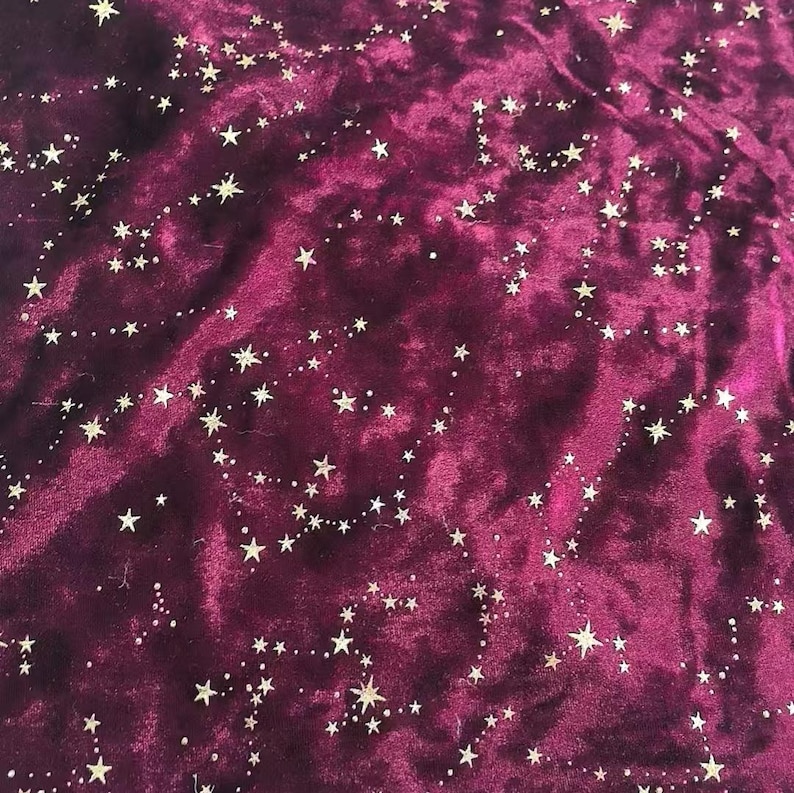 May include: Close-up of a deep burgundy velvet fabric with a constellation pattern. The design features small, gold-colored stars and connecting lines, creating a celestial theme. The fabric has a soft, textured appearance.