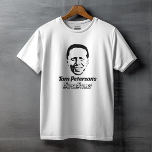 May include: White t-shirt with a black graphic of a man's face and the text "Tom Peterson's SuperStores". The shirt is hanging on a wooden hanger against a gray wall.